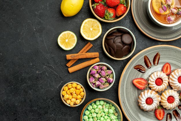 Top close-up view cookies with tea cookies with strawberry black tea with lemon bowls of chocolate and different sweets on the right side of table