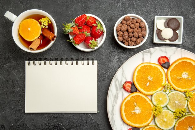 Top close-up view a cup of tea appetizing dish of citrus fruits and strawberries next to the bowls of chocolate strawberries and hizelnuts white notebook and cup of tea with lemon