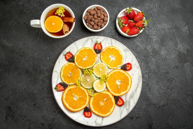 Top close-up view cup of tea and fruits plate of citrus fruits and chocolate-covered strawberries next to cup of tea with cinnamon and lemon plates of nuts and strawberries on the table