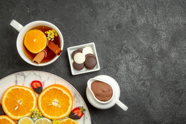 Top close-up view a cup of tea plate of citrus fruits and chocolate-covered strawberries next to the bowls of chocolate and chocolate cream and cup of tea with lemon and cinnabon sticks