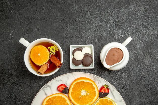 Top close-up view a cup of tea plate of citrus fruits and strawberries next to the bowls of chocolate and chocolate cream and cup of tea with lemon and cinnabon sticks