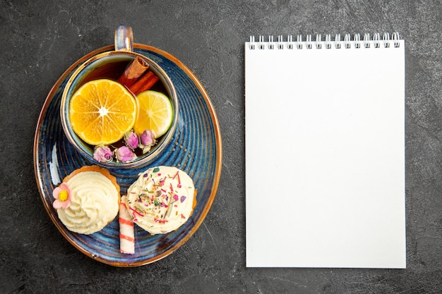 Top close-up view a cup of tea with lemon the appetizing cupcakes with cream next to the cup of herbal tea with lemon and cinnamon and white notebook on the dark table