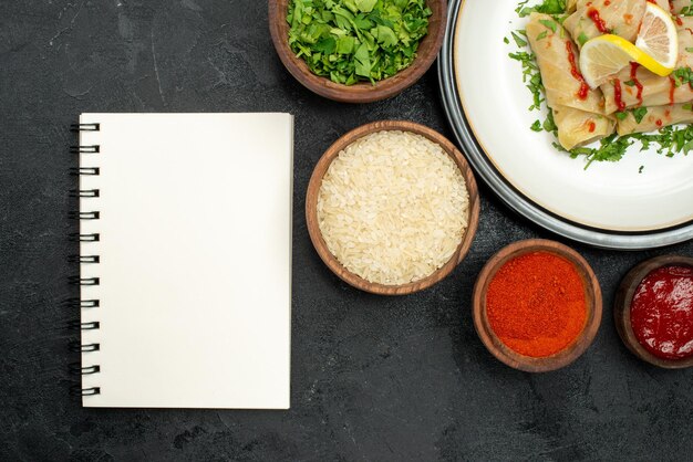 Top close-up view dish with sauce white plate of stuffed cabbage with lemon herbs and sauce and spices rice herbs and sauce in bowls next to white notebook on dark surface