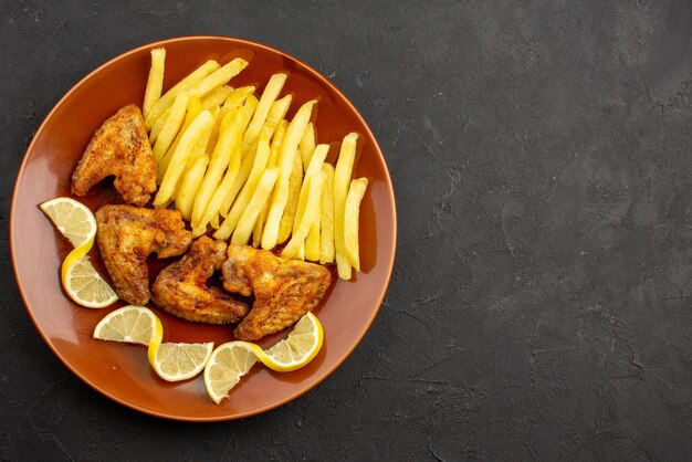 Top close-up view fastfood orange plate of appetizing french fries chicken wings and lemon on the left side of dark table