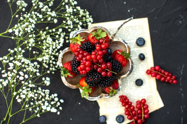 A top close up view fresh fruits colorful berries around paper and white flowers on the dark surface