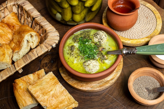 A top close up view hot soup inside brown round pot along with salted cucumbers bread sliced and spices