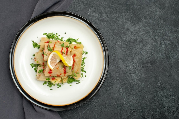 Free photo top close-up view plate on tablecloth white plate of stuffed cabbage with herbs lemon and sauce on grey tablecloth on the left side of dark table