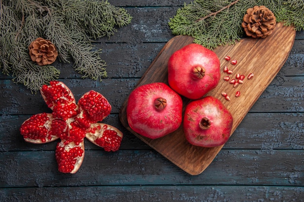 Free photo top close-up view pomegranate on board three ripe pomegranates on wooden board next to seeds of pomegranate spruce branches with cones and pilled pomegranate on table