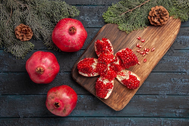 Free photo top close-up view pomegranate and branches pilled pomegranate on wooden board next to seeds of pomegranate spruce branches with cones knife and three pomegranates on grey table