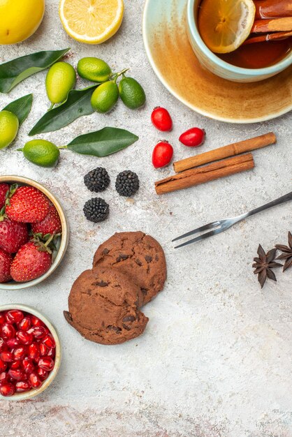 Free photo top close-up view strawberries cookies berries in bowls cookies a cup of tea lime and lemon cinnamon sticks fork on the table