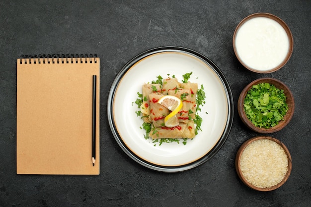 Top close-up view stuffed cabbage stuffed cabbage with herbs lemon and sauce on white plate and herbs rice and sour cream in plates next to cream notebook and pencil on black table