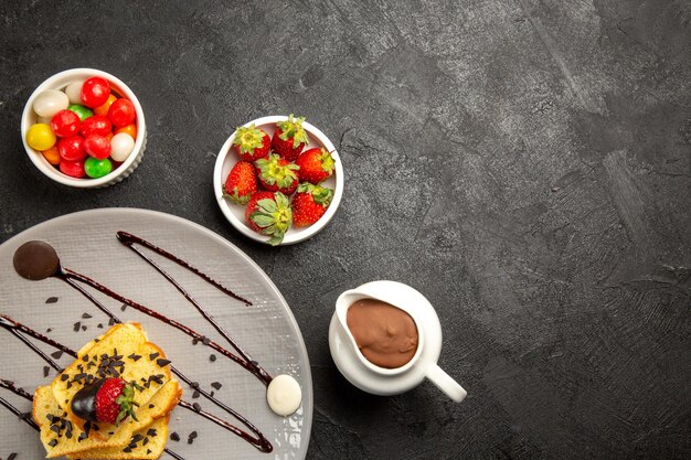 Top close-up view sweets bowls of sweets next to grey plate of pieces of cake with chocolate sauce and strawberries