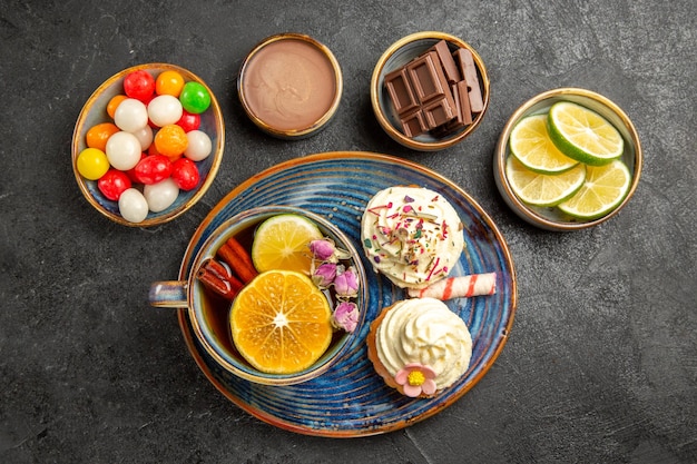 Top close-up view sweets on the table four bowls of chocolate candies and limes a plate of two cupcakes and the cup of herbal tea on the table