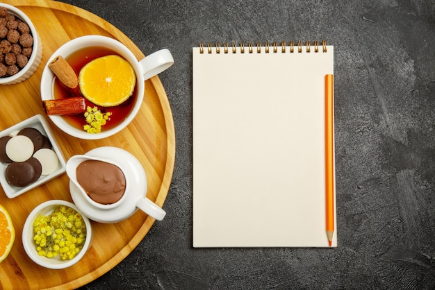 Top close-up view sweets on table notebook with yellow pencil next to the plate of chocolate berries lemon cinnamon sticks and a cup of tea with lemon