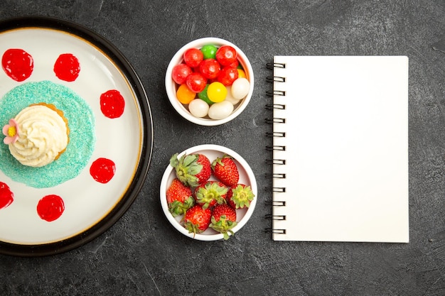 Top close-up view sweets on the table white notebook next to the plate of cupcake and bowls of sweets and strawberries on the black table