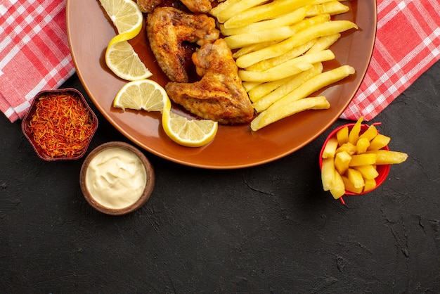 Top close-up view tasty chiken appetizing chicken wings french fries and lemon bowls of different types of sauces and spices in the center of the dark table