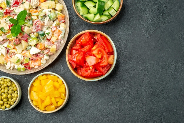 Top close view of bowl of vegetable salad with bowls of vegetables on side on dark green background