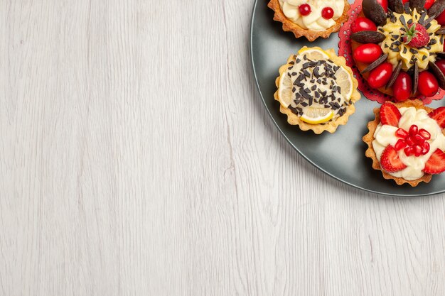 Top close view chocolate cake rounded with berry tarts in the grey plate on the top right of the white wooden table