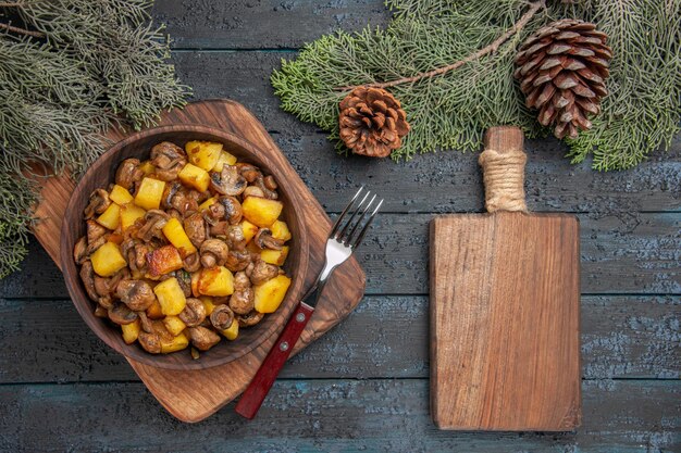 Top close view dish and cutting board wooden bowl of potatoes with mushrooms next to the cutting board and fork under spruce branches with cones