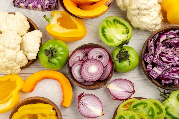 Top close view fresh vegetables cut green tomatoes cut red cabbage cut onion cut pumpkin cauliflower cut bell pepper in bowls on white wooden surface