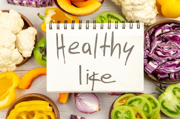 Top close view healthy life written on notebook cut green tomatoes cut red cabbage cut pumpkin cauliflower cut bell peppers in bowls on wooden surface