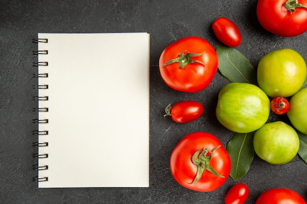 Free photo top close view red and green tomatoes bay leaves around a cherry tomato and a notebook on dark ground