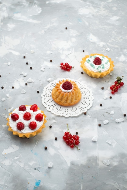 Top distant view little yummy cakes with cream and red fruits on the grey desk cake sweet biscuit sugar cream