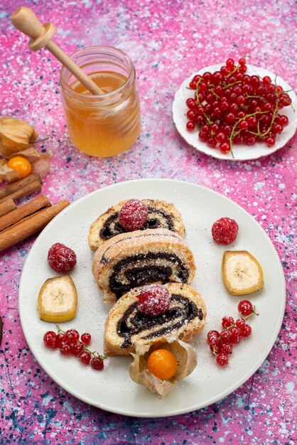 Top distant view roll cake slices with different fruits inside white plate with honey on the colored desk cake biscuit sweet fruit