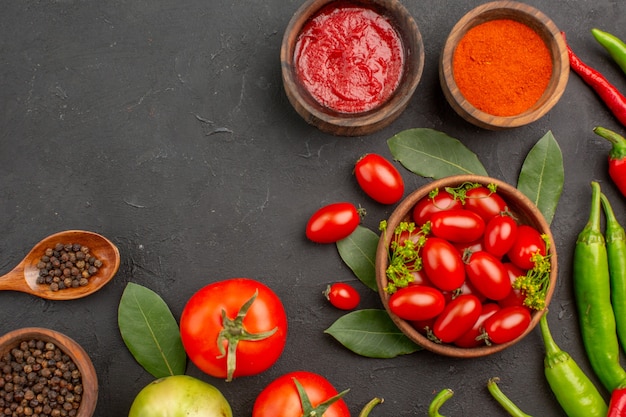 Top half view a bowl of cherry tomatoes hot red and green peppers and tomatos bay leaves spices in wooden spoons bowls of ketchup red pepper powder and black pepper on ground