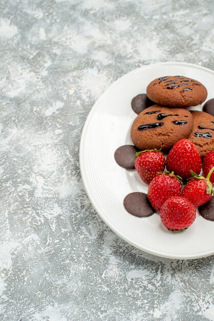 Top half view chocolate cookies strawberries and round chocolates on the white oval plate at the right side of the grey-white ground