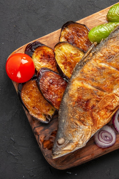 Top half view fish fry fried eggplants onion on wood serving board on dark surface