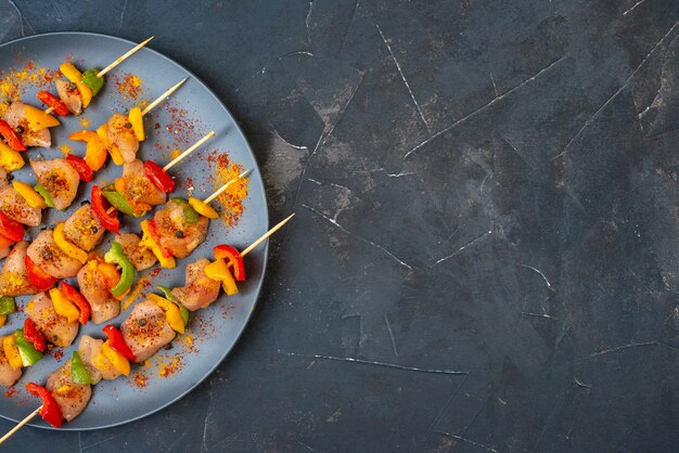 Top half view raw chicken skewers with spices on wooden board on dark