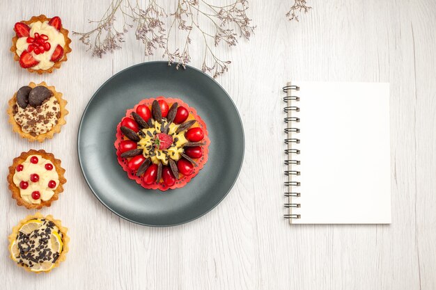 Top left side view berry cake in a grey plate and from top to the bottom different tarts and a notebookon the white wooden ground