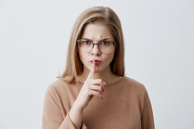 Top secret. Shot of cute blonde young female wearing eyeglasses, holding a finger on her lips, saying 'shh', asking to keep silent about her big secret, seriously looking at the camera