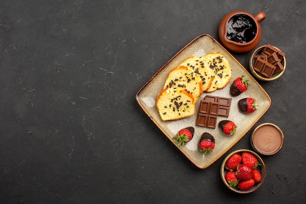 Free Photo top view appetizing cake cake with strawberries and chocolate between bowls of chocolate cream strawberries and chocolate on the right side of black table