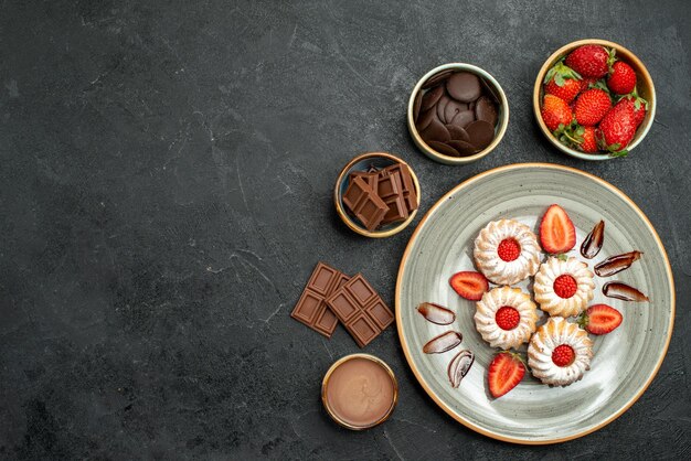 Top view appetizing sweets cookies with strawberry and chocolate sauce next to bowls of strawberry chocolate and sauce on dark table