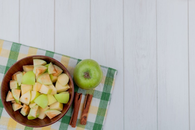 Free photo top view of apple cubes in bowl and whole one with cinnamon on plaid cloth and wooden background with copy space