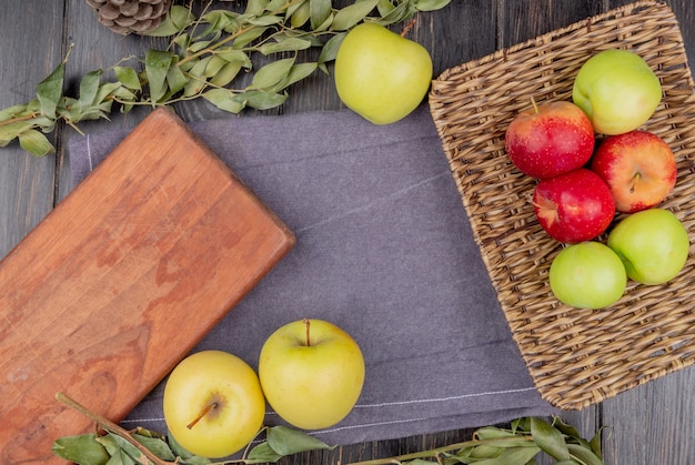 top view of apples in basket plate and on gray cloth with cutting board and leaves on wooden table