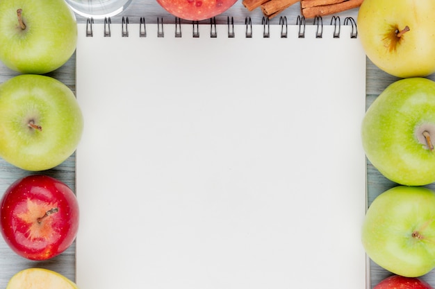 Top view of apples with cinnamon and water around note pad on wooden background with copy space