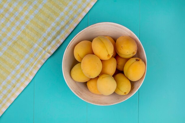 Top view of apricots in a bowl with a yellow checkered towel on a blue surface