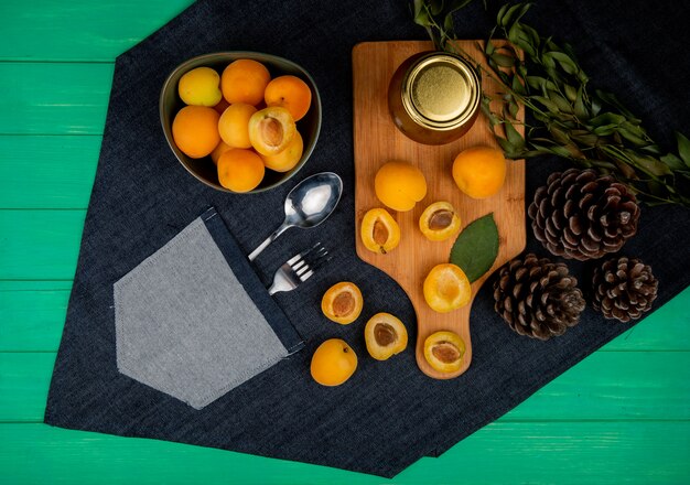 Top view of apricots and peach jam on cutting board with bowl of apricots pinecones leaves on jeans cloth with spoon and fork in pocket on green background