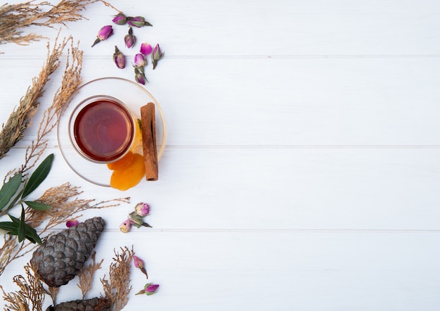 Top view of armudu glass of tea with dried apricots, cinnamon stick and dry rose buds scattered on white with copy space