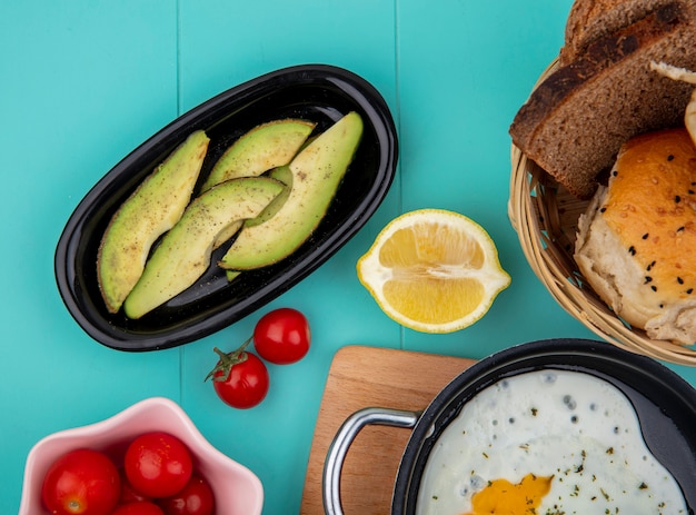 Top view of avocado slices on black plate with lemon tomato and with a bucket of breads on blue