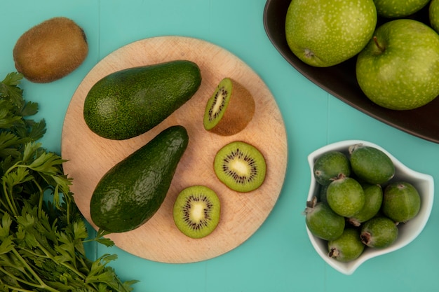 Top view of avocados with kiwi slices on a wooden kitchen board with feijoas on a bowl with apples on a bowl on a blue wall