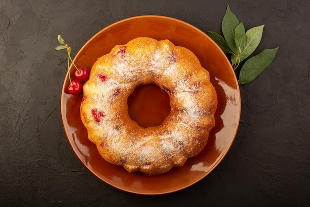 A top view baked fruit cake delicious round with red cherries inside and sugar powder inside round brown plate on dark