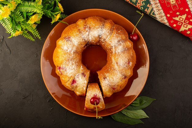 A top view baked fruit cake delicious round with red cherries inside and sugar powder inside round brown plate on dark