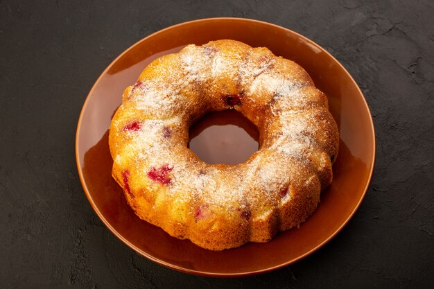 A top view baked fruit cake delicious round with red cherries inside and sugar powder inside round brown plate on dark