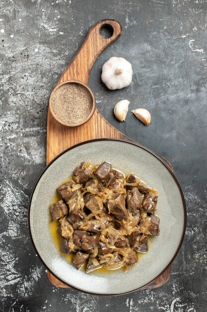 Top view baked liver and onion on plate black pepper on chopping board garlic on grey table