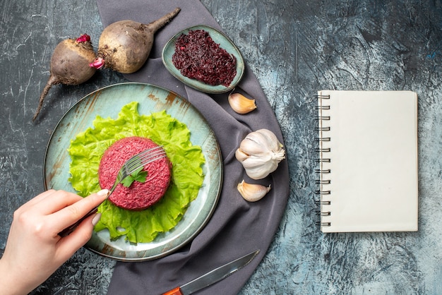 Top view beet salad on plate garlic beets grated beet in small bowl fork in female hand purple shawl notebook on grey table