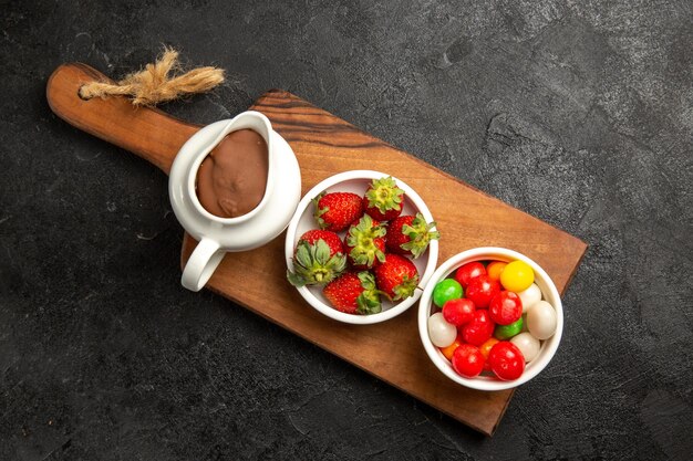 Top view berries bowls of chocolate sauce sweets and strawberries on the wooden cutting board on the dark table